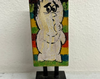 May include: A wooden figurine of a white character with a black hair and a rainbow striped background. The figurine is standing on a black wooden base with two black legs.