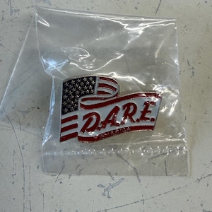 May include: A metal lapel pin enclosed in a clear plastic bag. The pin features a stylized American flag design with red, white, and blue enamel. The text D.A.R.E. is prominently displayed in red lettering, with the word AMERICA written below it.