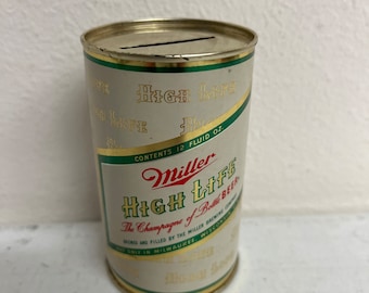 May include: A vintage metal Miller High Life beer can piggy bank. The can is gold and white with green accents. It says "Miller High Life, The Champagne of Bottle Beer" and "Brewed and Filled by the Miller Brewing Co. Only in Milwaukee, Wisconsin".