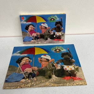 May include: A 100-piece jigsaw puzzle featuring Cabbage Patch Kids dolls at the beach. The dolls are wearing swimsuits and playing in the sand. There is a beach umbrella, a sandcastle, and a bucket and shovel.