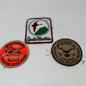 May include: Three embroidered patches: a rectangular white patch with a red bird and green mountain labeled 'Gender Mountain', a circular neon orange patch with a rifle and 'Wisconsin DNR Hunter Education', and a circular tan patch with a deer head.