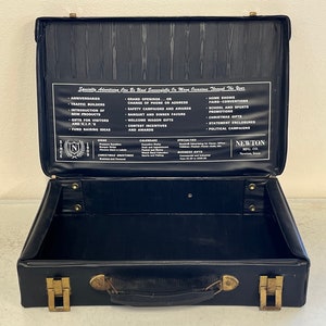 May include: Black vintage suitcase with a gold handle and clasps. The inside of the suitcase is lined with black fabric and has a white label with the text "Seminte Advertising Can Be Used Successfully On Many Occasions Throughout The Year." The label lists various occasions for which the suitcase could be used, such as anniversaries, traffic builders, grand openings, and more. The label also includes the text "Newton Mfg. Co. Newton, Iowa."