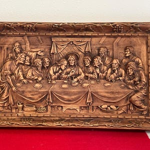 May include: A framed wood relief sculpture depicting the Last Supper. The scene shows Jesus and his disciples seated at a table, with a wooden background and a simple, ornate frame.