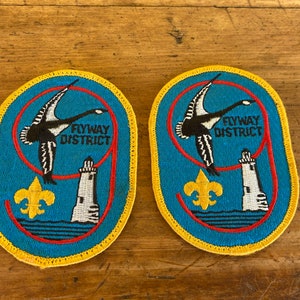 May include: Two blue and yellow embroidered patches with a red outline. The patches feature a black and white goose flying over a lighthouse and a yellow fleur-de-lis. The text "Flyway District" is embroidered in black on each patch.