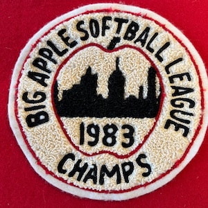 May include: A white and red embroidered patch with the text "Big Apple Softball League" and "1983 Champs" around a black silhouette of the New York City skyline.