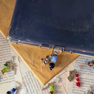 Könnte beinhalten: Eine Sammlung kleiner, dekorativer Anhänger zum Anstecken an eine Tablethülle. Jeder Anhänger besteht aus einer einzigartigen Metallfigur, wie einem Wolf oder einer Fee, kombiniert mit bunten Glasperlen in Blau-, Grün- und Rottönen.