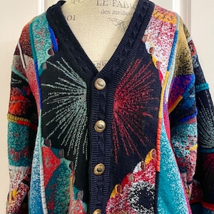 May include: A vibrant, button-up cardigan with a V-neck. The sweater has a patchwork design in shades of blue, red, yellow, and teal. Navy blue trim and cuffs, with gold buttons. A striking piece of knitwear.
