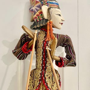 May include: An Indonesian Wayang Golek puppet. The puppet features a painted face, elaborate headdress, and a detailed costume with gold and red detailing. It is controlled by wooden rods.