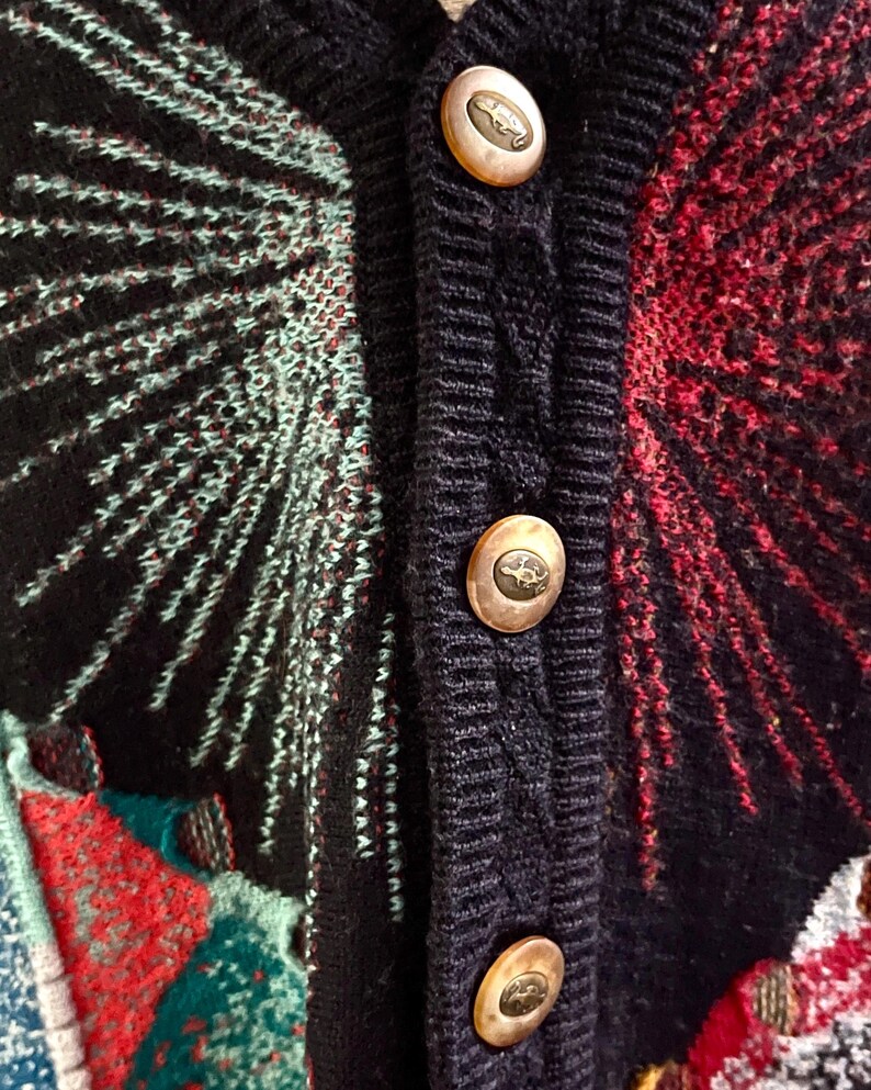 Puede incluir: Primer plano de un c&aacute;rdigan vintage con fondo azul marino y patrones coloridos. El su&eacute;ter presenta botones dorados con un peque&ntilde;o dise&ntilde;o de animal. El dise&ntilde;o incluye detalles en rojo, verde azulado y blanco.