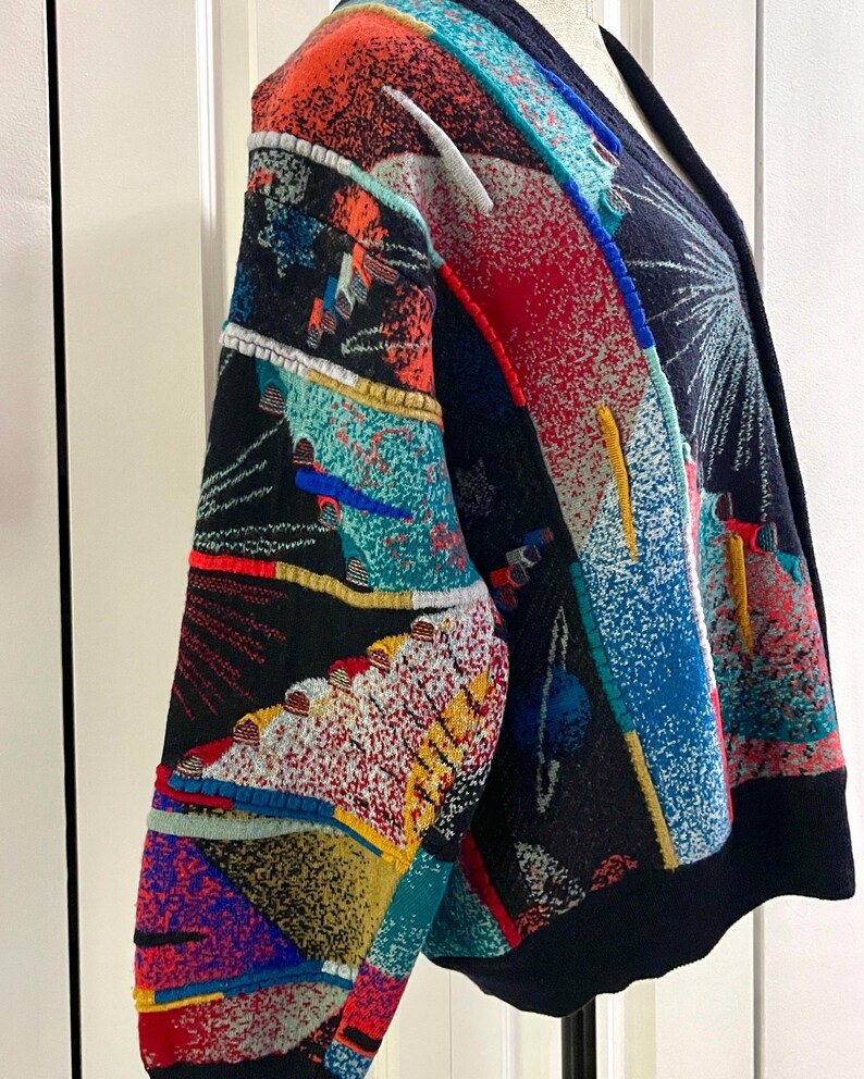 Puede incluir: Un c&aacute;rdigan estampado y colorido con base negra. El dise&ntilde;o incorpora formas geom&eacute;tricas y patrones abstractos en rojo, azul, naranja y turquesa. El c&aacute;rdigan tiene cuello en V y mangas largas.