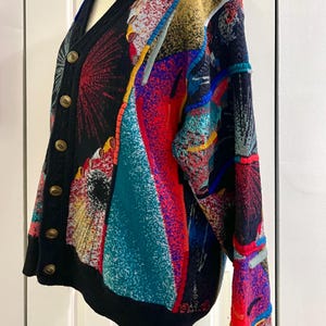 Puede incluir: Un c&aacute;rdigan multicolor con cuello en V y botones dorados. El su&eacute;ter presenta una base negra con patrones abstractos vibrantes en rojo, azul, amarillo y turquesa. La prenda se exhibe en un maniqu&iacute;. El dise&ntilde;o recuerda a una pintura abstracta.