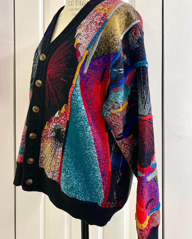 Puede incluir: Un c&aacute;rdigan multicolor con cuello en V y botones dorados. El su&eacute;ter presenta una base negra con patrones abstractos vibrantes en rojo, azul, amarillo y turquesa. La prenda se exhibe en un maniqu&iacute;. El dise&ntilde;o recuerda a una pintura abstracta.