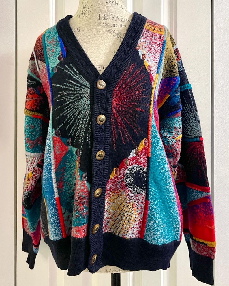 May include: A vibrant, button-up cardigan with a V-neck. The sweater has a patchwork design in shades of blue, red, yellow, and teal. Navy blue trim and cuffs, with gold buttons. A striking piece of knitwear.