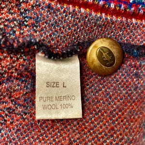 Puede incluir: Primer plano de una colorida tela de punto con una etiqueta que dice "SIZE L. PURE MERINO WOOL 100%". Se ve un bot&oacute;n dorado con un dise&ntilde;o de lagarto. El patr&oacute;n de punto presenta tonos rojos, azules y verdes.