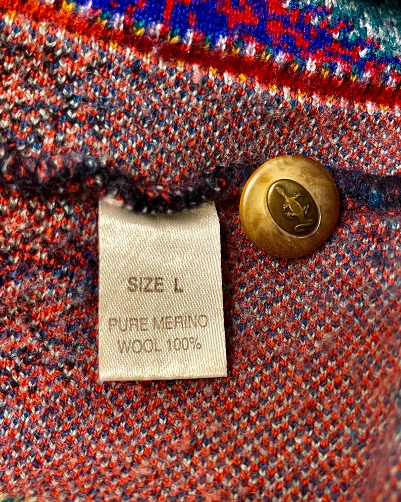 Puede incluir: Primer plano de una colorida tela de punto con una etiqueta que dice "SIZE L. PURE MERINO WOOL 100%". Se ve un bot&oacute;n dorado con un dise&ntilde;o de lagarto. El patr&oacute;n de punto presenta tonos rojos, azules y verdes.