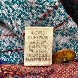 Puede incluir: Primer plano de una prenda de punto con una etiqueta que dice "HAND WASH IN LUKE WARM WATER. LAY FLAT TO DRY. WARM IRON. OR DRY CLEAN. MADE IN AUSTRALIA." La prenda tiene patrones turquesa, blancos, rojos y negros y un bot&oacute;n marr&oacute;n.
