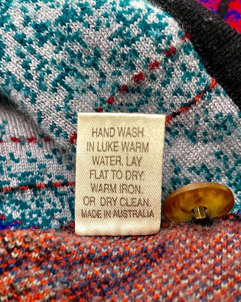 Puede incluir: Primer plano de una prenda de punto con una etiqueta que dice "HAND WASH IN LUKE WARM WATER. LAY FLAT TO DRY. WARM IRON. OR DRY CLEAN. MADE IN AUSTRALIA." La prenda tiene patrones turquesa, blancos, rojos y negros y un bot&oacute;n marr&oacute;n.