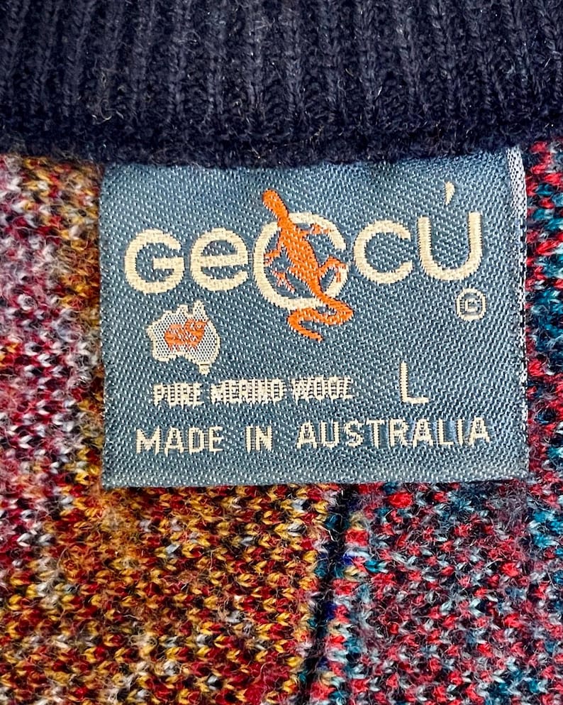 Puede incluir: Primer plano de una etiqueta azul con la marca "geccu" y un logotipo de canguro, cosida a una prenda de lana colorida. La etiqueta dice "PURE MERINO WOOL" y "MADE IN AUSTRALIA". La prenda es una mezcla de rojo, amarillo y azul.
