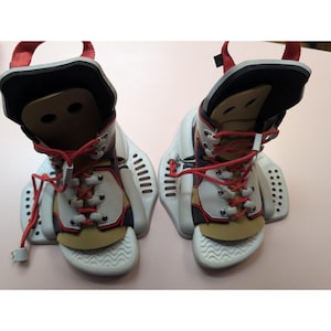May include: Pair of white, black, red, and tan wakeboard boots with red laces and straps. The boots have a high-top design with a molded footbed and are designed to attach to a wakeboard. The boots are shown on a pink surface.