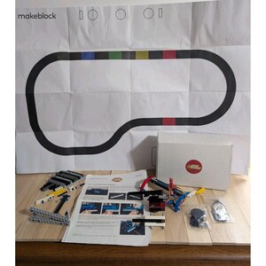 May include: A Makeblock robotics kit featuring a large white paper track with a black oval path, a white cardboard box with a circular logo, a printed instruction manual, various grey plastic building beams, and small electronic components.