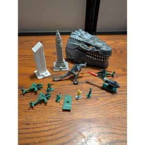 Könnte beinhalten: Eine Sammlung von Spielzeugfiguren und -strukturen, darunter ein grauer Godzilla-Kopf, ein Dinosaurier, zwei Wolkenkratzer, grüne Hubschrauber, Miniatursoldaten und eine Kanone. Das Spielzeug ist auf einer Holzoberfläche angeordnet.