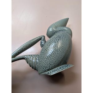 May include: A ceramic watering can with a speckled, gray-green finish. The spout is long and pointed, and the handle curves gracefully. The base is flat and wide. The watering can is designed for indoor or outdoor use.