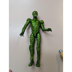 May include: A metallic green action figure of a humanoid character with a detailed, textured design. The figure has a helmet, armor-like plating, and articulated limbs. The figure is standing on a neutral surface.
