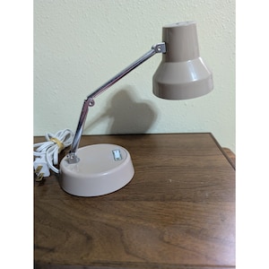 May include: A beige desk lamp with a chrome adjustable arm and a matching base. The lamp has a conical shade and a power cord with a standard US plug. The lamp is on a wooden surface.