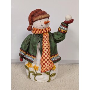 May include: A decorative snowman figurine with a carrot nose, wearing a red hat, green jacket, and a scarf with a yellow and red checkered pattern. The snowman is holding a snowball in its right hand. Yellow flowers decorate the base.