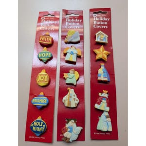 May include: Three red card strips with Christmas-themed button covers. The buttons feature religious words and images like angels, stars, and a nativity scene. The text "Holiday Button Covers" is at the top of each card. The words "FAITH", "HOPE", "JOY", "PROMISE", and "HOLY NIGHT" are visible.