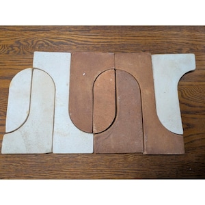 May include: A set of four interlocking ceramic tiles featuring a smooth, matte finish. Two tiles are a light cream colour, and two tiles are a warm terracotta brown. Each tile has a curved, arch-like shape that fits together to form a pattern.