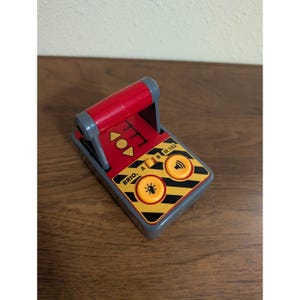 May include: A red and gray toy remote control with the text "BRIO A IR 102". The remote has two yellow buttons with black accents and a red handle. The remote is on a wooden surface.