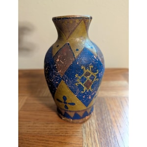 Puede incluir: Un pequeño jarrón pintado a mano con un cuerpo bulboso y un cuello estrecho. El jarrón presenta un patrón de arlequín de diamantes azules, marrones y amarillos, con un emblema decorativo y detalles florales. El jarrón mide unos 15 cm de alto.