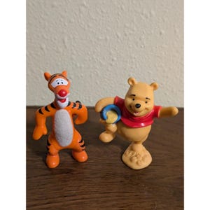 Puede incluir: Dos figuras de personajes de Winnie the Pooh. Tigger, un tigre naranja con rayas negras, está de pie junto a Winnie the Pooh, un oso amarillo que lleva una camisa roja y sostiene un objeto azul y amarillo. Las figuras están sobre una superficie de madera.