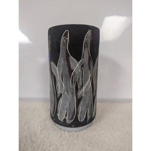 Puede incluir: Un jarrón cilíndrico negro con base blanca. El jarrón presenta un diseño abstracto gris y blanco con detalles en rojo. El diseño parece ser de figuras estilizadas. El jarrón es probablemente de cerámica o piedra.