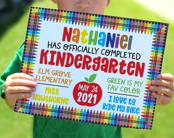 May include: A colorful sign with a crayon border announcing that Nathaniel has officially completed kindergarten. The sign includes the name of the school, Elm Grove Elementary, the teacher's name, Miss Hawthorne, and the date May 24, 2021. The sign also includes a green apple with a red stem and leaf, and the text "Green is my fav color" and "I love to ride my bike".