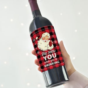 May include: A dark wine bottle with a label featuring Santa Claus in a red and black plaid design. The label reads "He sees YOU when you're DRINKING". The bottle is held against a white background with soft, out-of-focus lights.