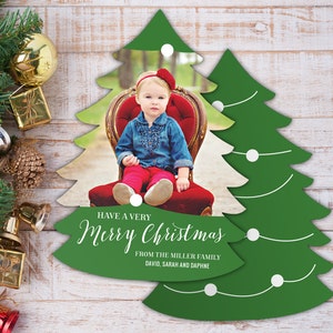 May include: A green Christmas tree-shaped card with a white border and white lights. The card has a photo of a young girl wearing a red headband and a blue shirt sitting on a red chair. The text on the card reads "Have a Very Merry Christmas from the Miller Family David, Sarah and Daphne".