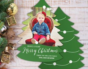 May include: A green Christmas tree-shaped card with a white border and white lights. The card has a photo of a young girl wearing a red headband and a blue shirt sitting on a red chair. The text on the card reads "Have a Very Merry Christmas from the Miller Family David, Sarah and Daphne".