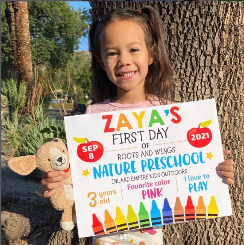 May include: A girl is holding a sign that reads "Zaya's First Day of Roots and Wings Nature Preschool". The sign also includes the date "September 8th", the year "2021", and the words "Inland Empire Kids Outdoors", "Favourite Colour Pink", and "I love to Play". There is a row of colourful crayons at the bottom of the sign.
