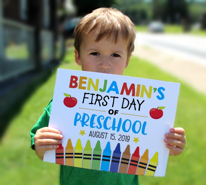 Puede incluir: Un cartel blanco con el texto "Benjamin's First Day of Preschool" en letras de colores. El cartel tambi&eacute;n tiene dos manzanas rojas y una fila de l&aacute;pices de colores. La fecha "August 15, 2019" est&aacute; impresa debajo del texto.