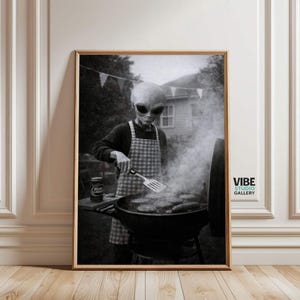 Puede incluir: Impresión en blanco y negro enmarcada de un extraterrestre asando hamburguesas en una parrilla de carbón. El extraterrestre lleva un delantal a cuadros y sostiene una espátula. Una lata de cerveza está sobre una mesa. Vibe Studio Gallery.