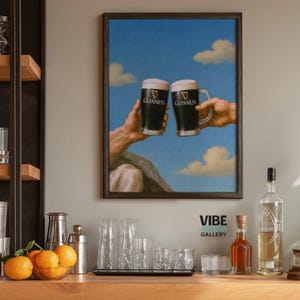 May include: Framed art print featuring two hands holding Guinness beer mugs against a blue sky with clouds. The Guinness logo is visible on the mugs. The print is displayed in a dark wooden frame. The text "VIBE STUDIO GALLERY" is at the bottom.