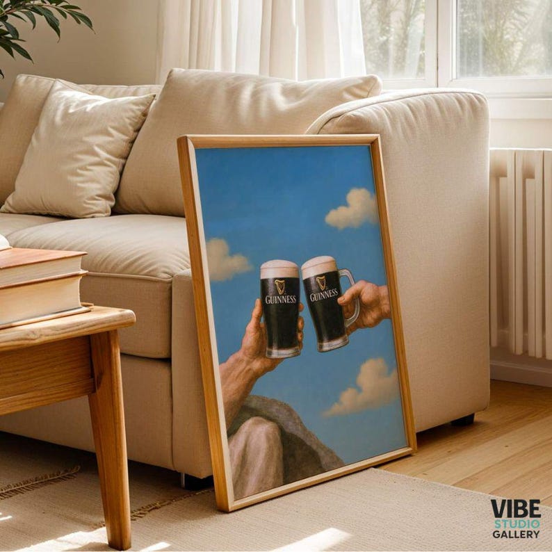 May include: A framed print featuring two Guinness beer mugs held up against a blue sky with clouds. The print has a wooden frame and the Guinness logo is visible on the mugs. The artwork is displayed in a living room setting.