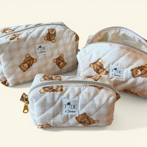 May include: Three quilted cosmetic bags with a teddy bear pattern. The bags are in varying sizes and feature a cream and tan gingham pattern with brown teddy bear illustrations. Each bag has a zipper closure and a label that reads "Tide & Twine."