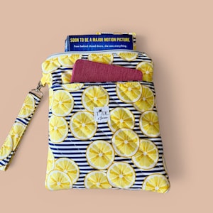 May include: A quilted pouch with a lemon slice pattern on a navy blue and white striped background. The pouch has a zipper closure and a wrist strap. A book with the text "SOON TO BE A MAJOR MOTION PICTURE" is visible.