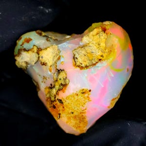 Puede incluir: Un ópalo en bruto con forma de corazón que presenta una base blanca lechosa y translúcida con destellos vibrantes de rosa neón, naranja y amarillo, intercalados con parches de roca matriz rugosa y texturizada de color tostado.