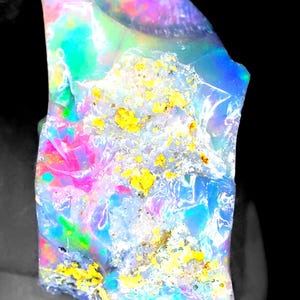 May include: A raw, uncut opal gemstone featuring a translucent, iridescent body with vibrant flashes of neon blue, green, and hot pink. The surface displays a textured, crystalline structure with embedded patches of bright yellow mineral inclusions.