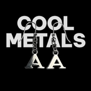 May include: Silver-toned letter "A" earrings with chain accents, hanging from curved ear wires. The earrings are set against a black background with the white text "COOL METALS".