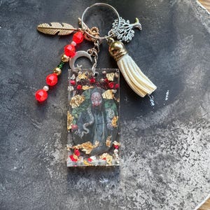 May include: A rectangular resin keychain featuring an embedded image of a masked figure, gold leaf flakes, and red beads. It includes a silver-tone metal ring with a gold feather charm, a silver tree charm, a crescent moon charm, and a white tassel.